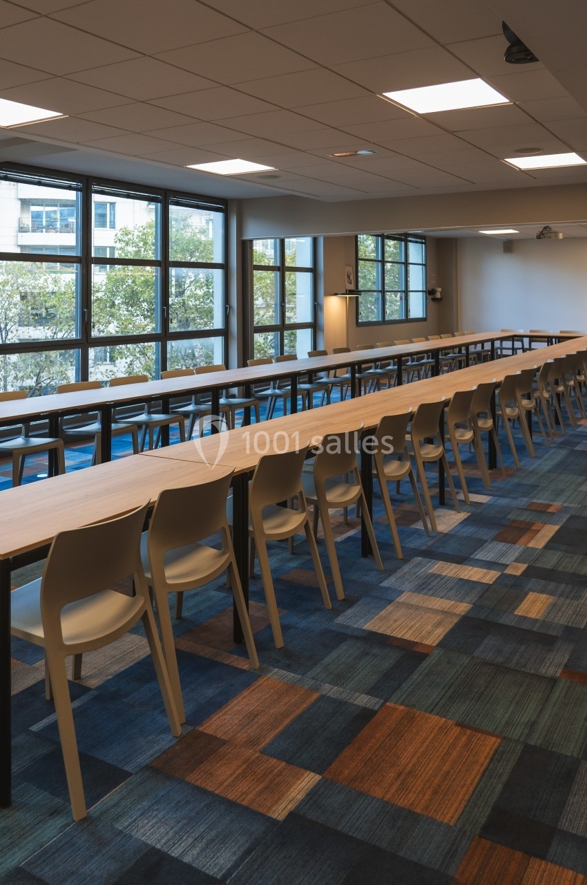 Salle de réunion lumineuse avec de longues tables en bois, des chaises alignées et un sol à motifs colorés.