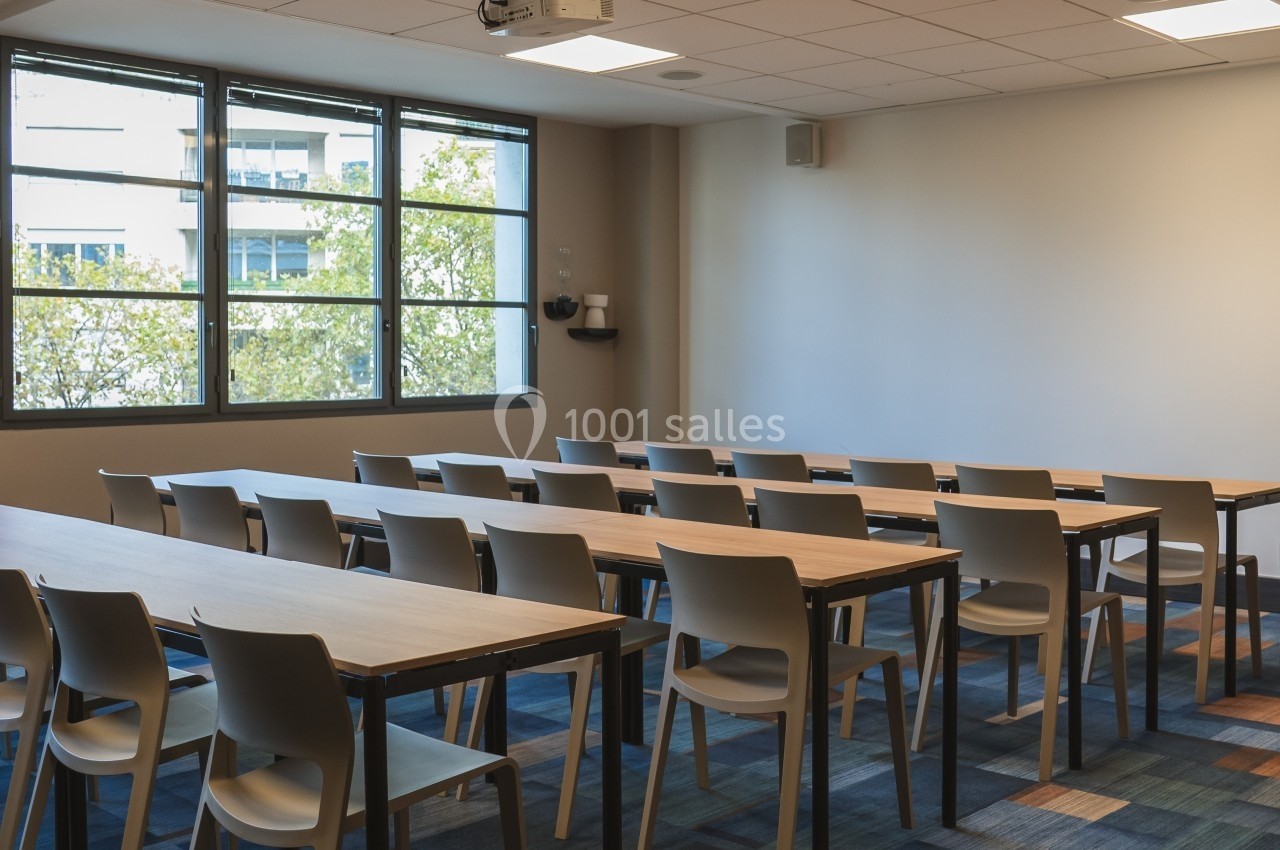 Salle de réunion lumineuse avec des tables et des chaises alignées face à un mur blanc et un projecteur au plafond.