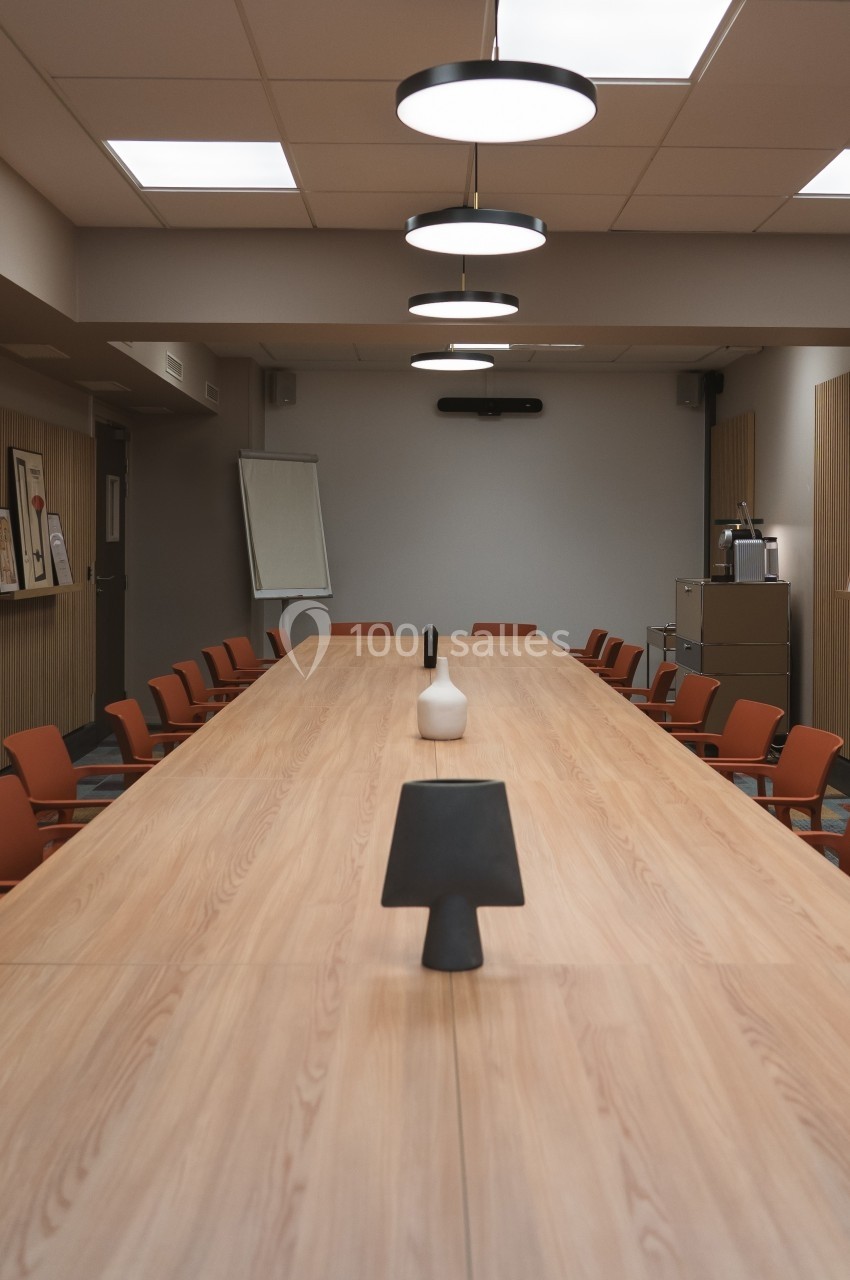 Salle de réunion avec une grande table en bois, des chaises orange, un tableau blanc et des cadres accrochés aux murs.