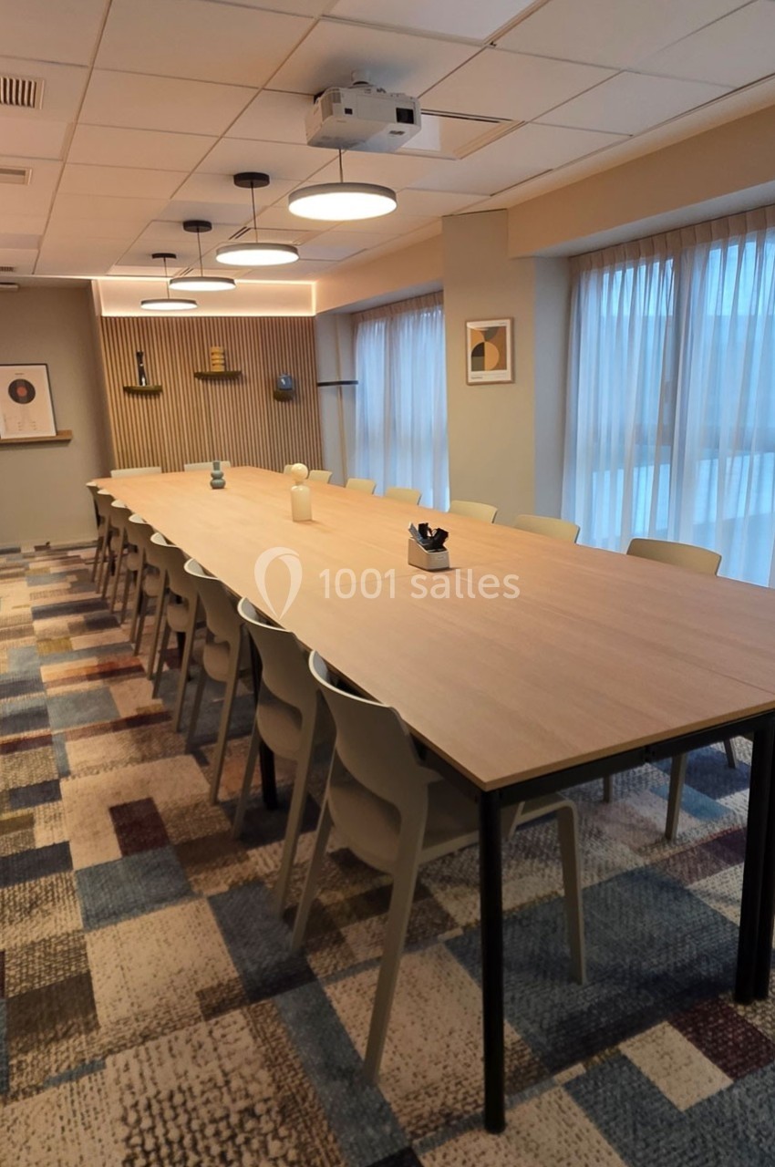 Salle de réunion lumineuse avec une grande table rectangulaire entourée de chaises, éclairée par des suspensions modernes.