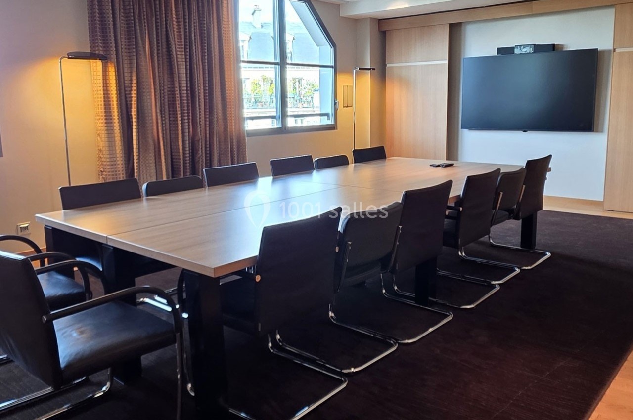 Salle de réunion moderne avec une grande table rectangulaire, des chaises noires et un écran mural.
