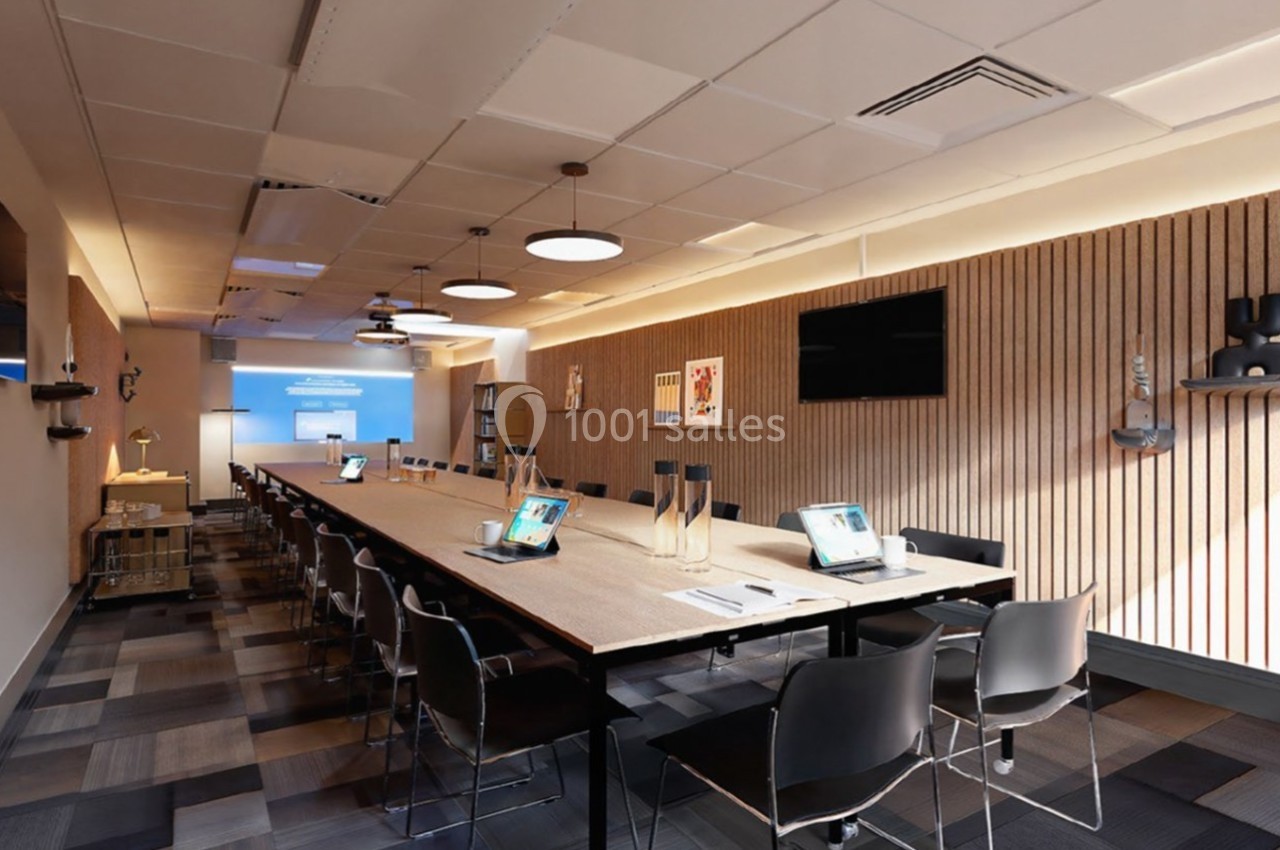 Salle de réunion moderne avec une grande table, des chaises, des ordinateurs portables et un écran de projection allumé.