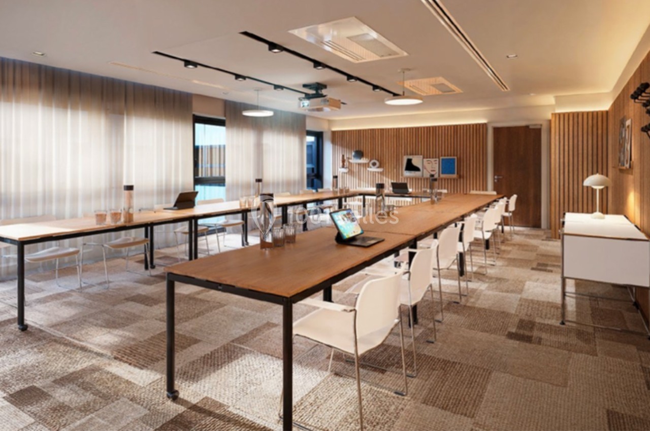 Salle de réunion moderne avec tables en bois, chaises blanches, éclairage naturel et équipements audiovisuels.