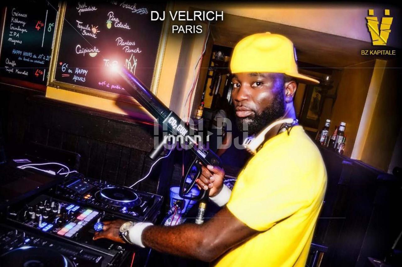 Un DJ portant une casquette jaune mixe sur des platines dans un bar avec des menus écrits à la craie en arrière-plan.