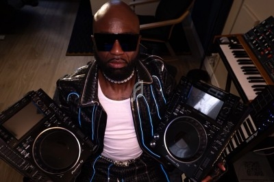 Un DJ en tenue originale mixe en extérieur devant une foule assise à des tables de café en terrasse.