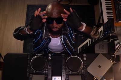 Un DJ en tenue originale mixe en extérieur devant une foule assise à des tables de café en terrasse.
