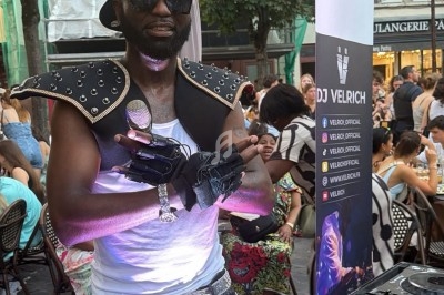 Un DJ en tenue originale mixe en extérieur devant une foule assise à des tables de café en terrasse.
