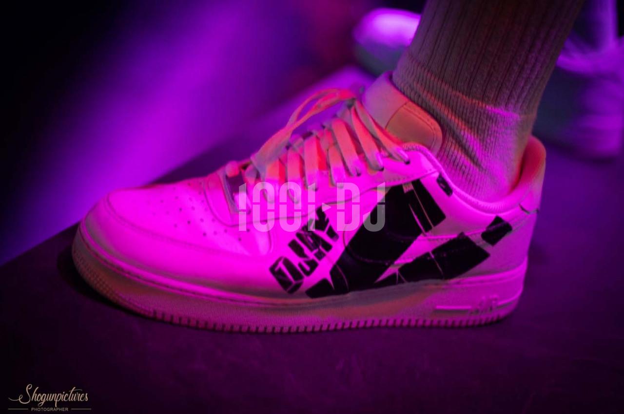 Une chaussure de sport blanche avec des motifs noirs, éclairée par une lumière violette, portée avec une chaussette blanche.