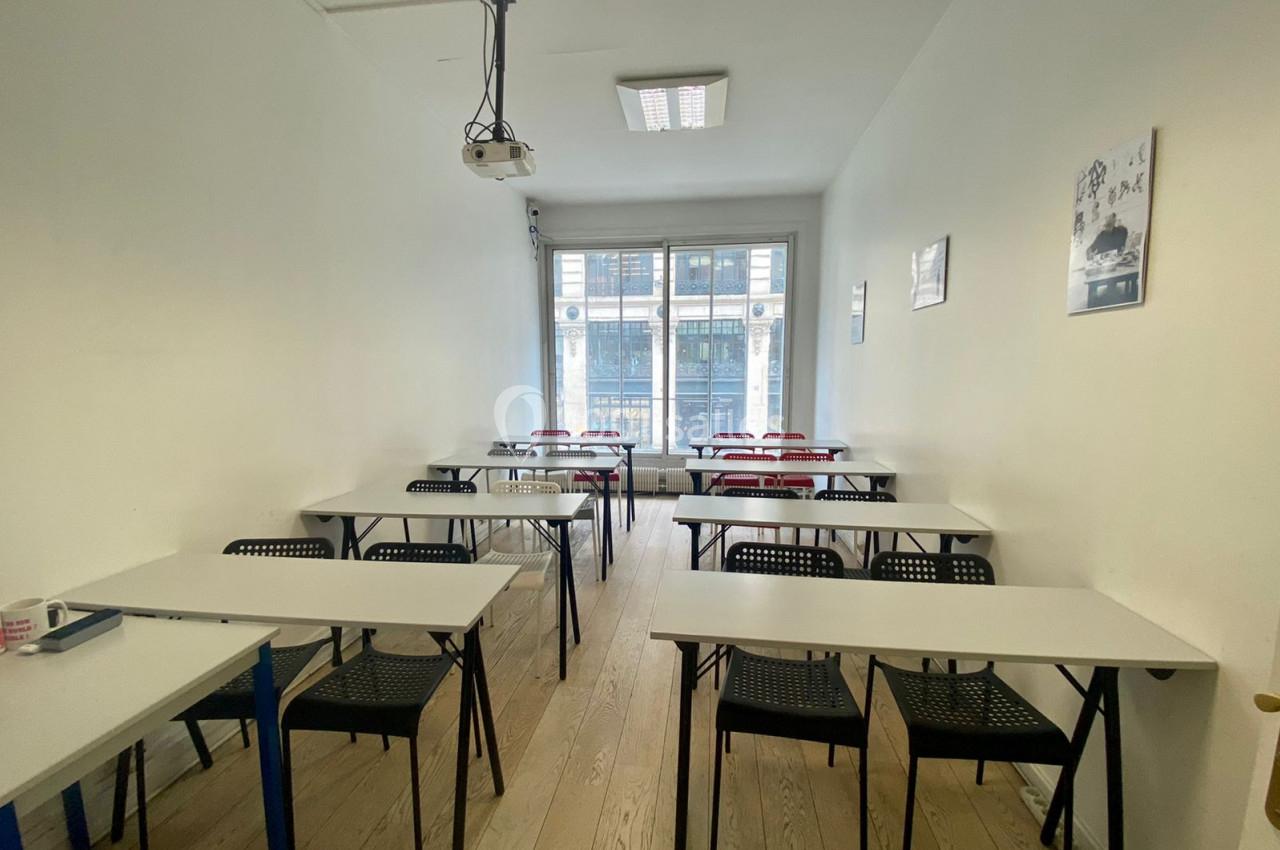 Salle de classe lumineuse avec des tables blanches, des chaises noires et un projecteur au plafond.