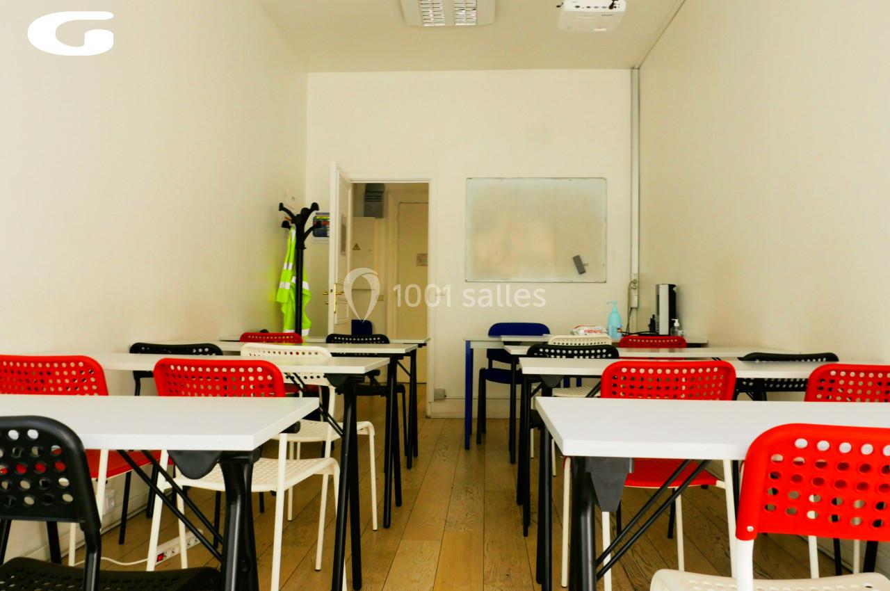 Salle de classe lumineuse avec des tables blanches, chaises rouges et noires, tableau blanc et projecteur au plafond.