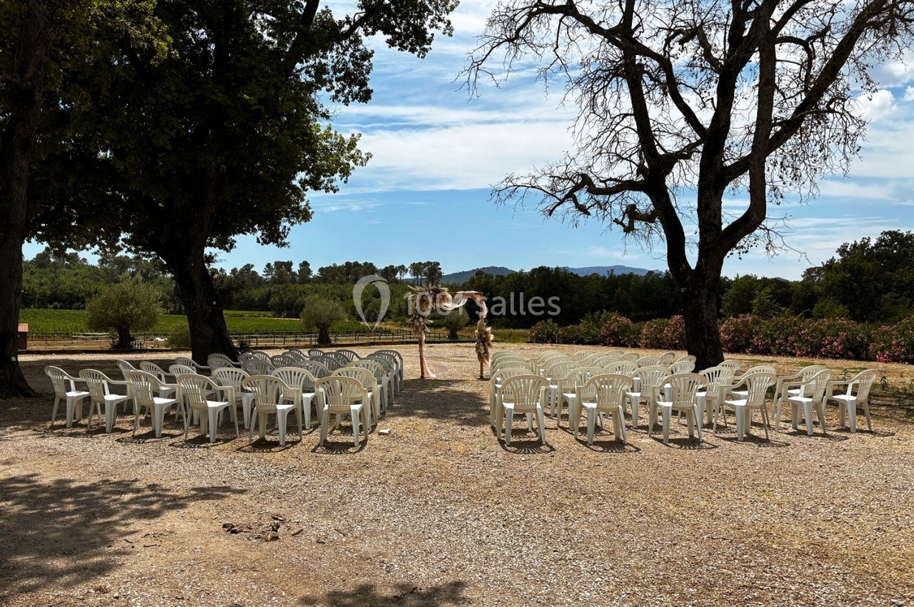 Chaises blanches disposées en rangées en plein air, face à une arche décorée, entourées d'arbres et de végétation.