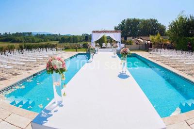 Allée centrale bordée de chaises blanches menant à une arche décorée de fleurs, dans un cadre extérieur arboré.