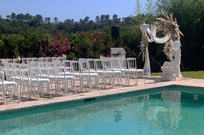 Allée centrale bordée de chaises blanches menant à une arche décorée de fleurs, dans un cadre extérieur arboré.