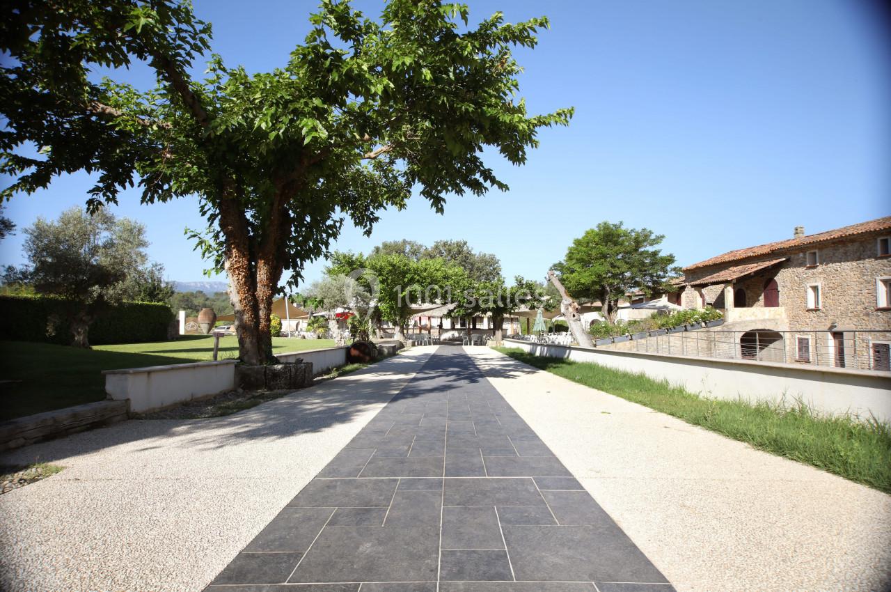 Allée pavée bordée d'arbres et de pelouses, menant à des bâtiments en pierre sous un ciel dégagé.