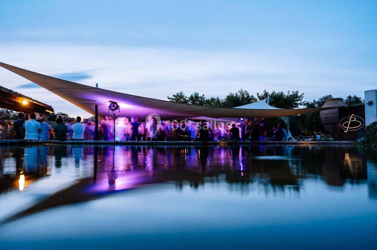Vue d'une soirée en extérieur avec éclairages violets, reflet dans l'eau et structure tendue au-dessus des invités.