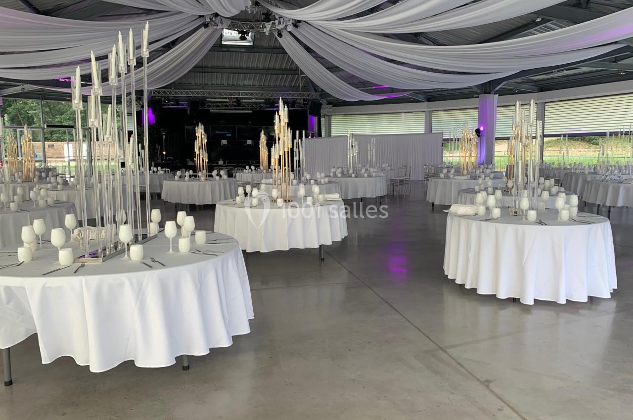Salle de réception décorée avec des tables rondes, nappes blanches, chandeliers et drapés suspendus au plafond.