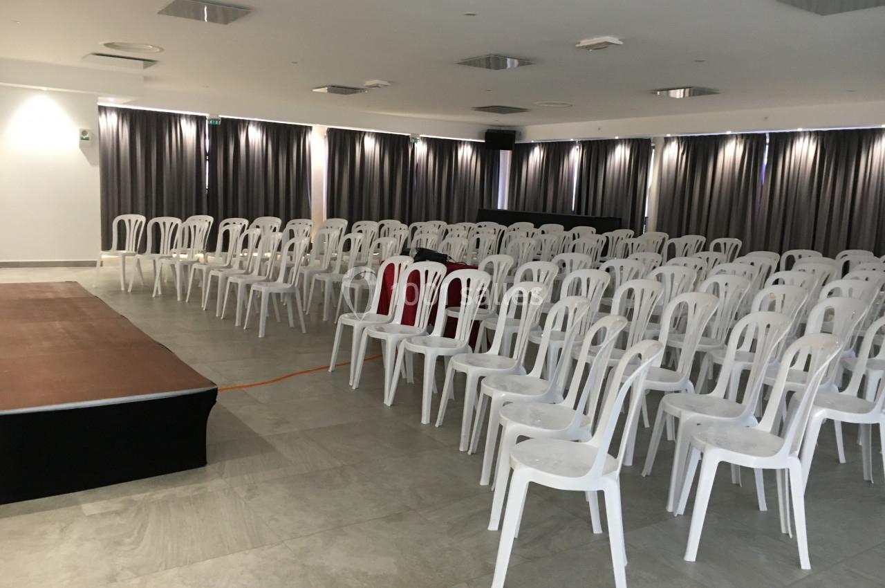 Salle de conférence vide avec des rangées de chaises blanches en plastique et une petite scène à gauche.