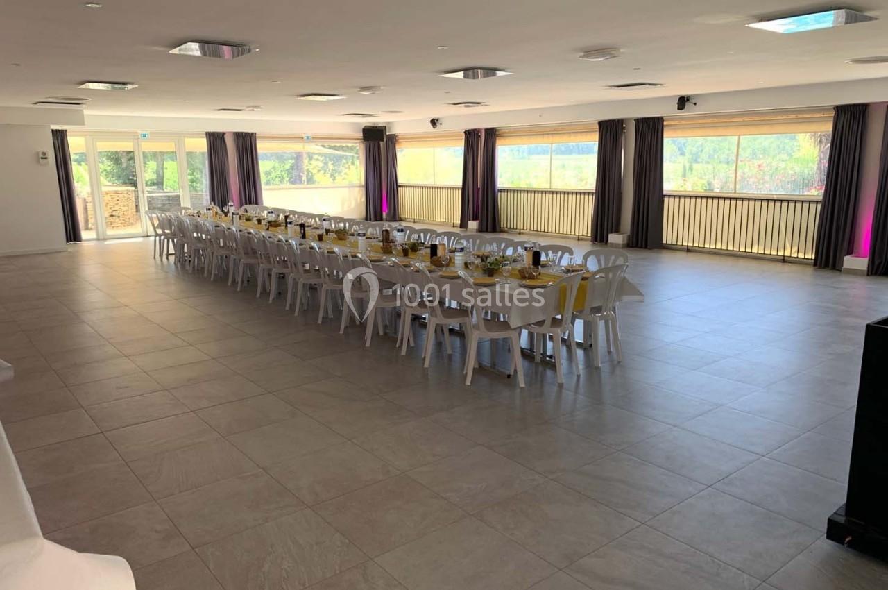 Grande salle lumineuse avec de longues tables dressées pour un repas, entourée de baies vitrées donnant sur l'extérieur.