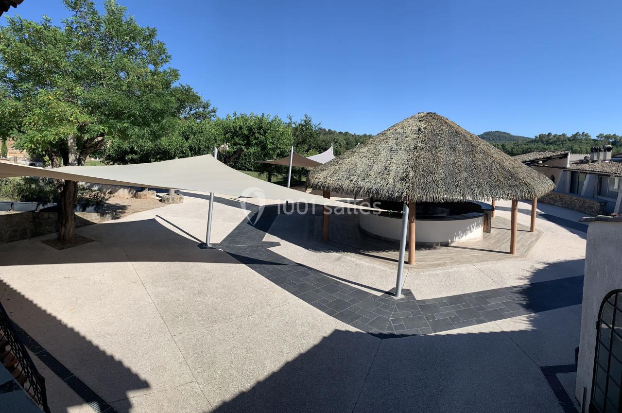 Espace extérieur avec pergola en chaume, voiles d'ombrage et sol en béton, entouré de végétation et bâtiments.