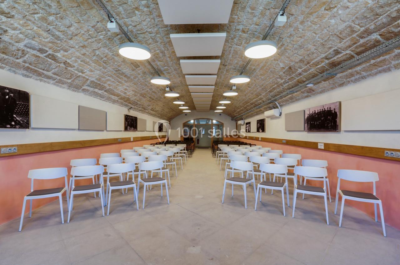 Salle voûtée en pierre avec rangées de chaises blanches alignées face à une porte et des panneaux muraux.