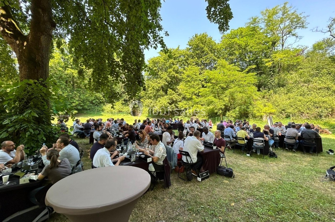 Groupe de personnes assises à des tables rondes en plein air, dans un cadre verdoyant et ensoleillé.