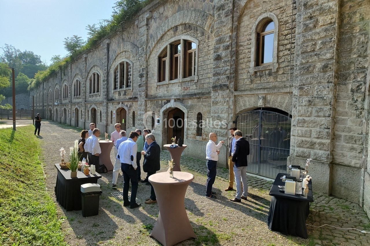 Groupe de personnes discutant debout autour de tables hautes devant un bâtiment en pierre avec des arches.