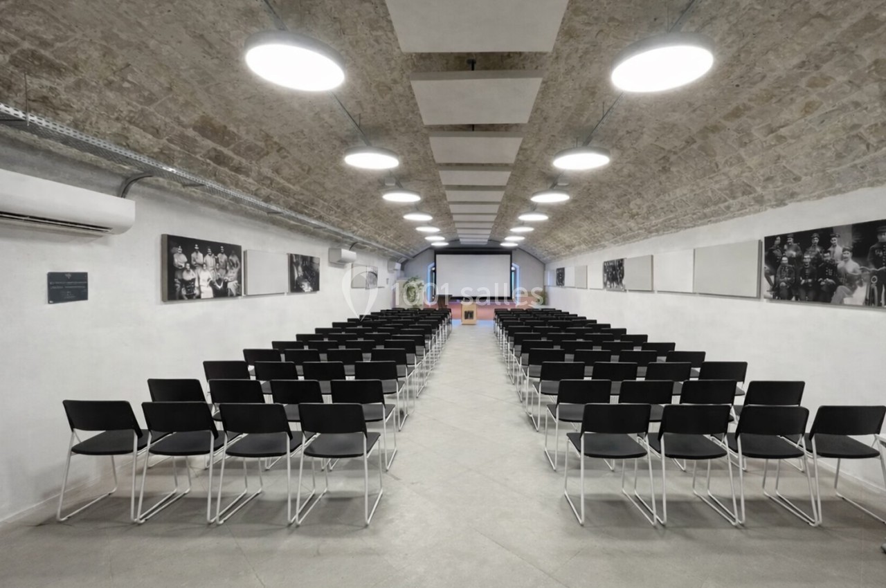 Salle de conférence avec rangées de chaises noires alignées face à un écran de projection sous un plafond voûté.