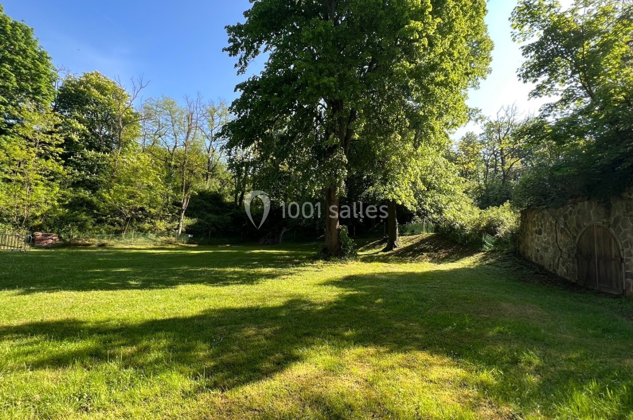 Pelouse verdoyante entourée d'arbres sous un ciel bleu, avec un mur en pierre et une porte en bois à droite.