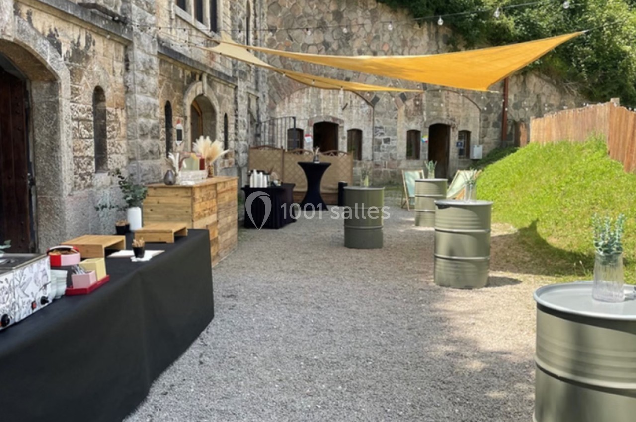 Cour extérieure avec tables en tonneaux, nappes noires, décorations naturelles et voiles d'ombrage près d'un bâtiment en…