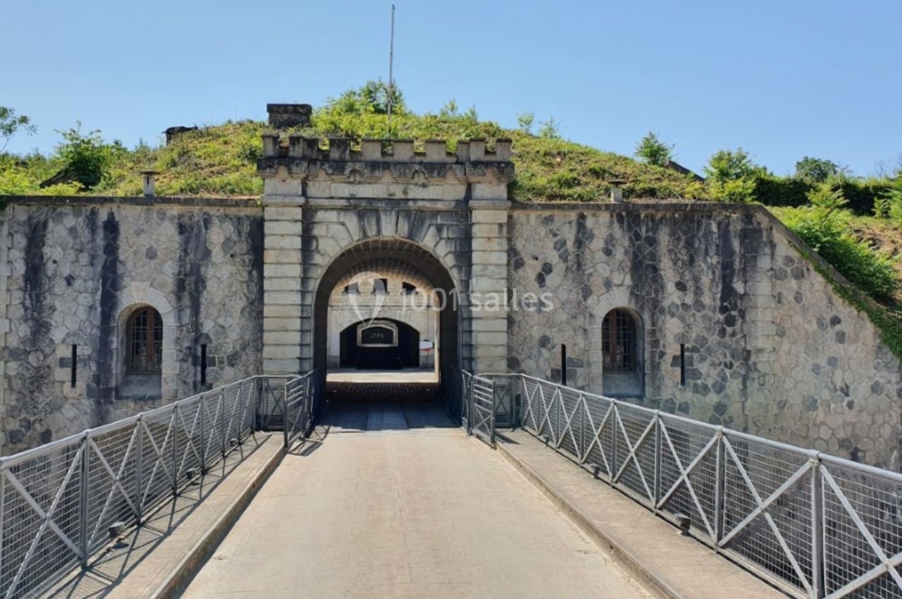 Entrée d'un fort en pierre avec un pont métallique, entouré de végétation.