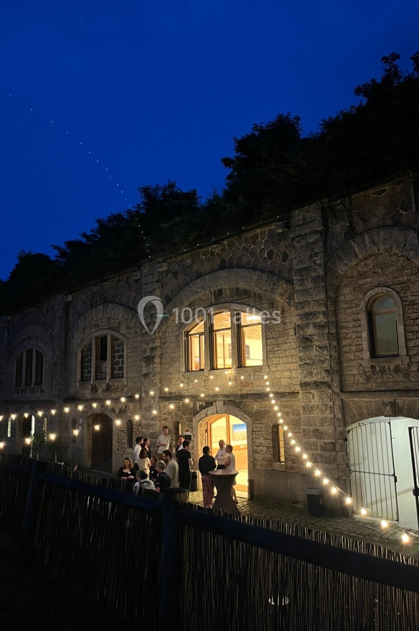 Façade en pierre éclairée par des guirlandes lumineuses, avec un groupe de personnes discutant à l'extérieur au crépuscule.