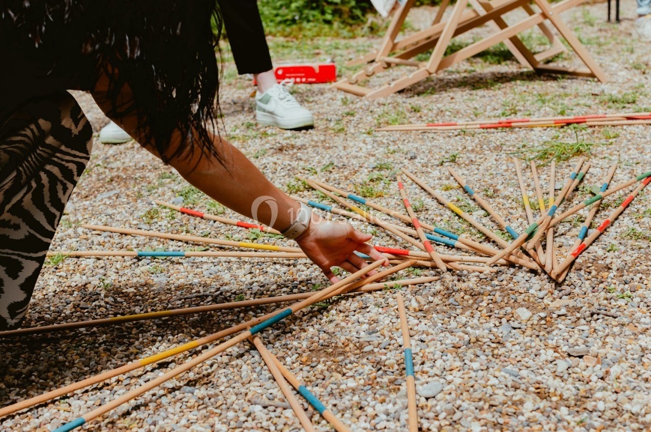 Personne ramassant des baguettes de mikado en bois coloré éparpillées sur un sol de gravier.