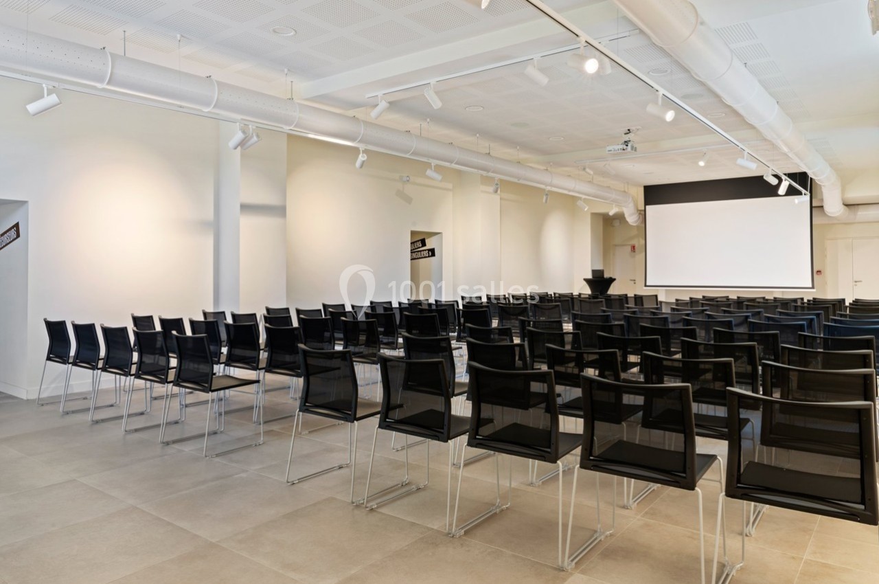 Salle de conférence lumineuse avec des rangées de chaises noires alignées face à un écran de projection blanc.