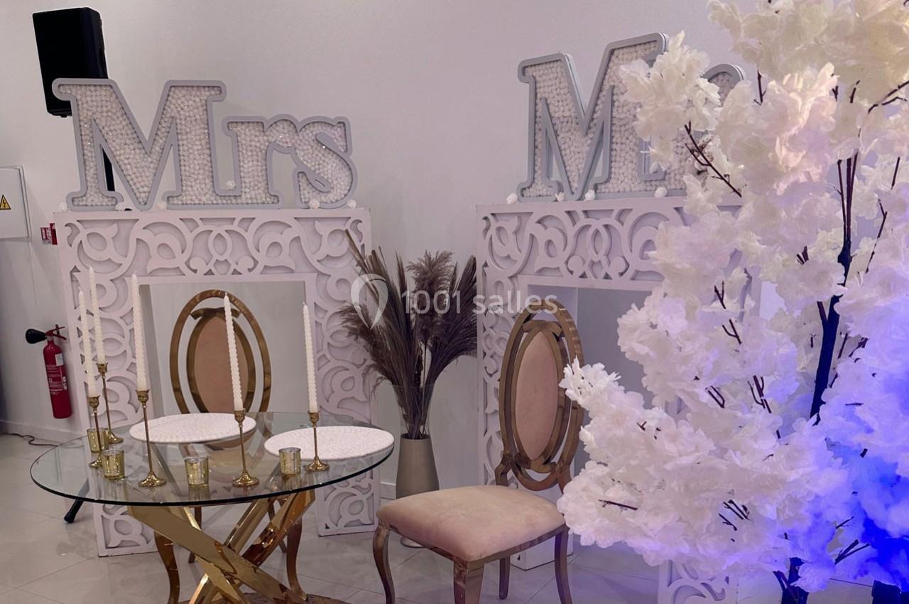 Décor de mariage avec chaises élégantes, table en verre, lettres ’Mrs’ et arbre artificiel blanc illuminé.