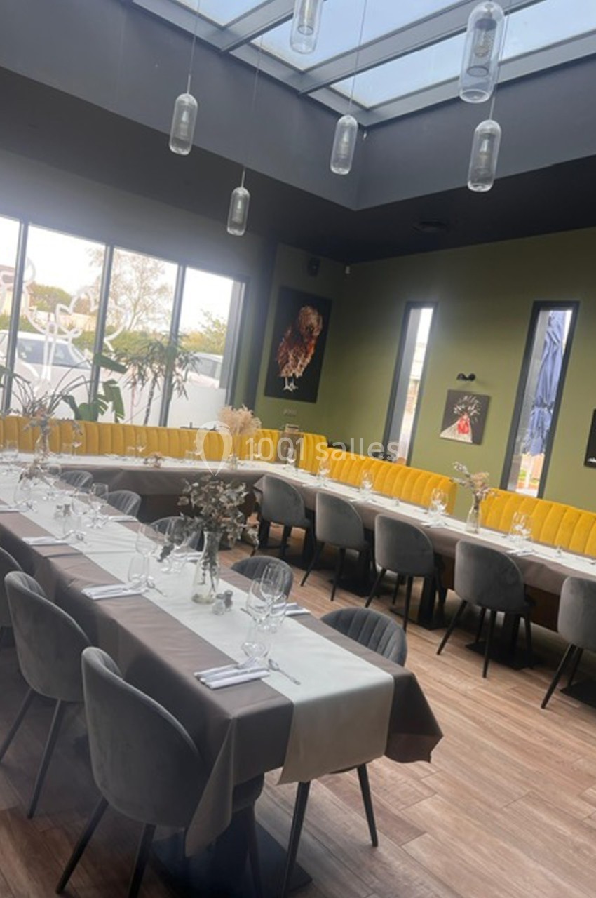 Salle de restaurant lumineuse avec de grandes baies vitrées, tables dressées et banquettes jaunes le long du mur.