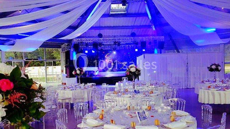 Salle de réception décorée avec des tables dressées, des chaises transparentes et un éclairage coloré sur une scène.