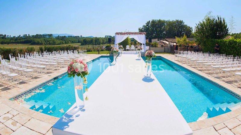 Allée blanche décorée de fleurs menant à une arche de mariage, entourée d'une piscine et de chaises disposées en extérieur.
