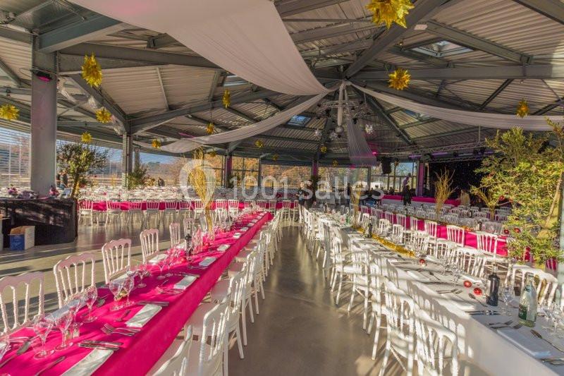 Salle de réception lumineuse avec de longues tables dressées, nappes roses et décorations suspendues dorées.