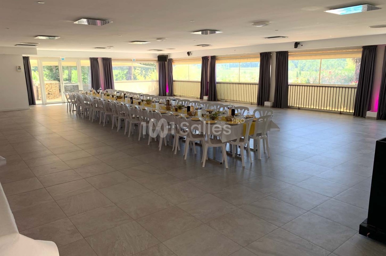 Grande salle lumineuse avec de longues tables dressées pour un repas, entourée de baies vitrées donnant sur l'extérieur.