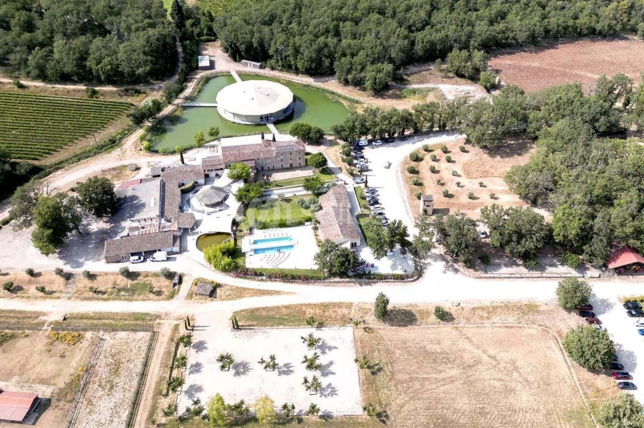 Vue aérienne d'un domaine avec bâtiments en pierre, piscine, étang, parking et végétation environnante.