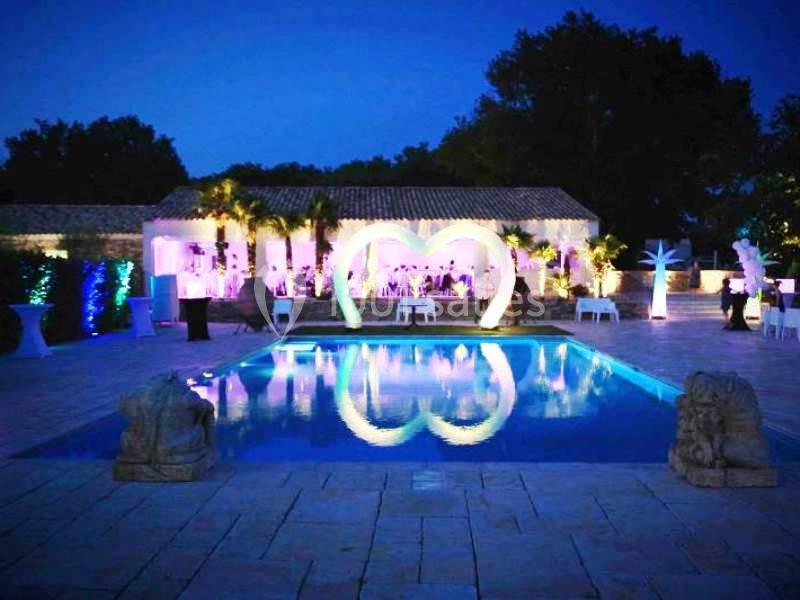 Piscine éclairée en soirée avec une arche lumineuse en forme de cœur et une salle décorée en arrière-plan.