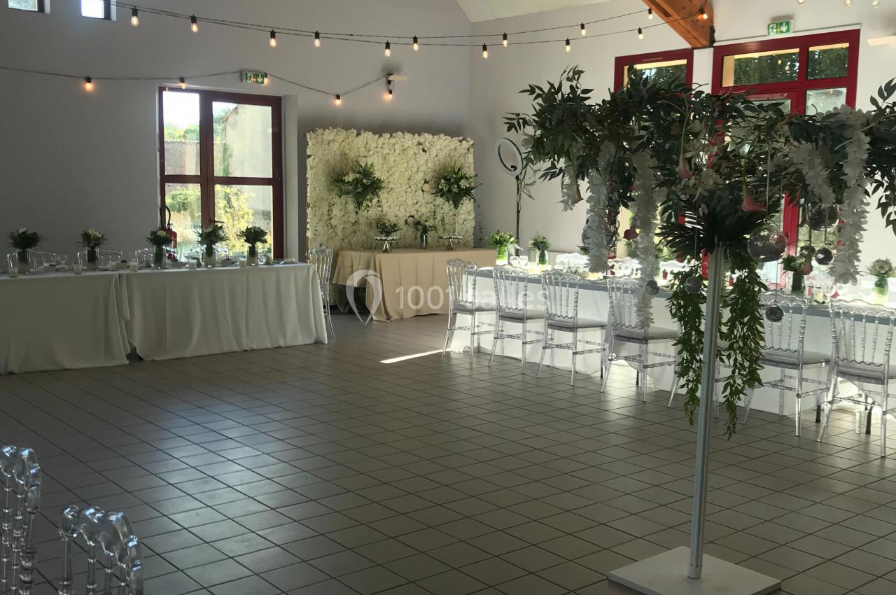 Salle de réception décorée pour un événement, avec tables dressées, chaises transparentes et guirlandes lumineuses…