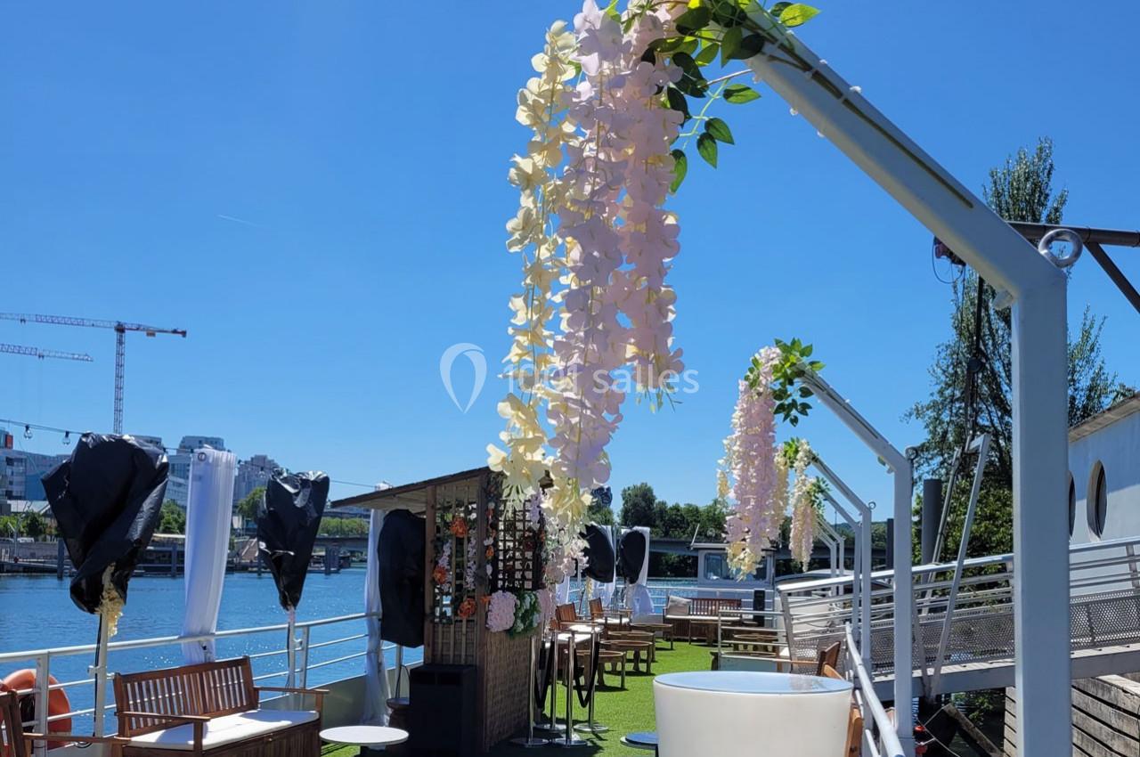 Terrasse aménagée sur une péniche avec mobilier en bois, décorations florales suspendues et vue sur une rivière.