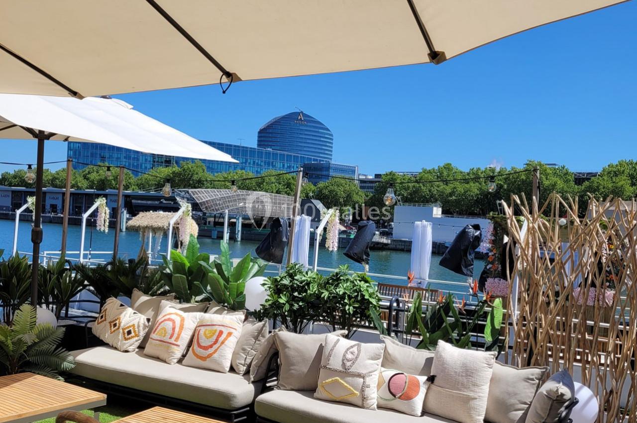 Terrasse aménagée avec canapés, coussins colorés, plantes et vue sur une rivière bordée de verdure et bâtiments modernes.