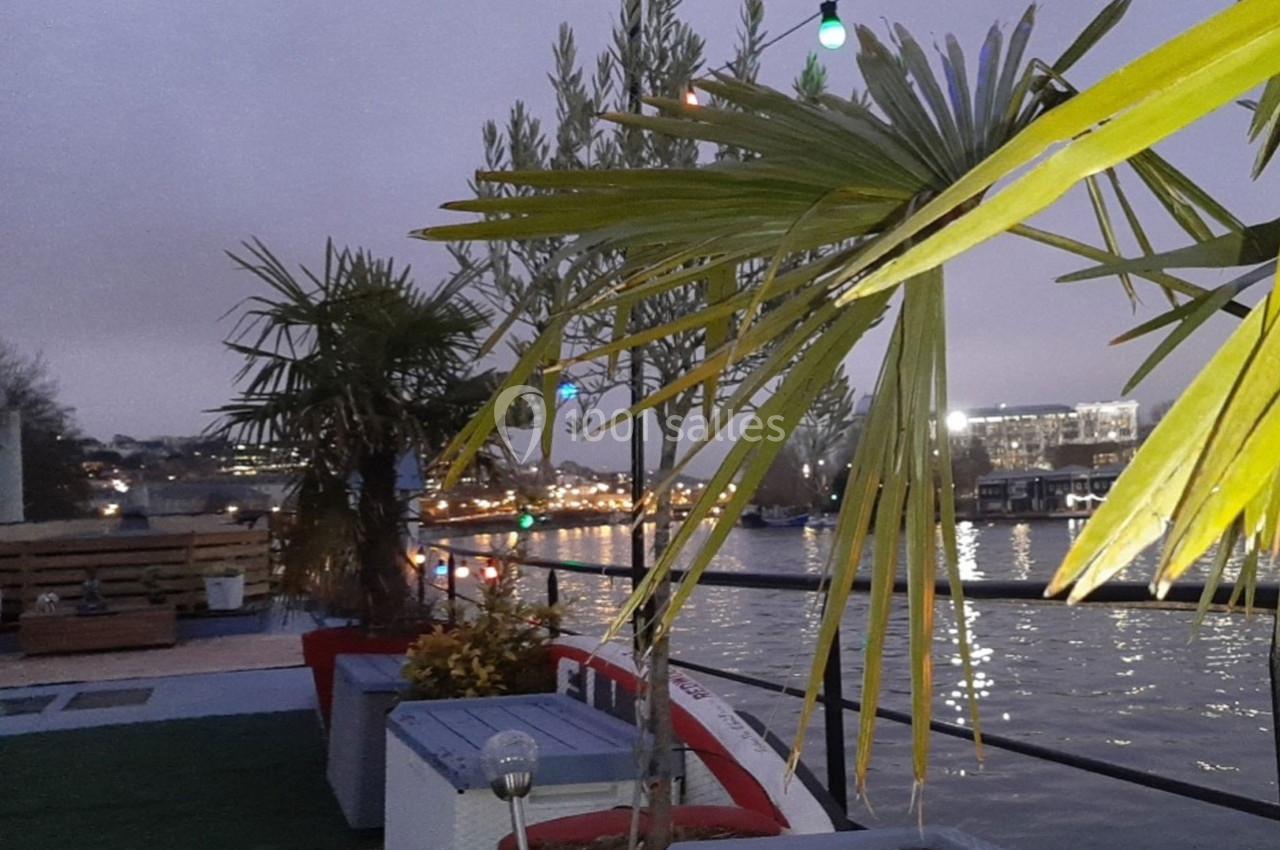 Terrasse au bord de l'eau avec des palmiers, des guirlandes lumineuses et une vue sur des bâtiments éclairés au loin.