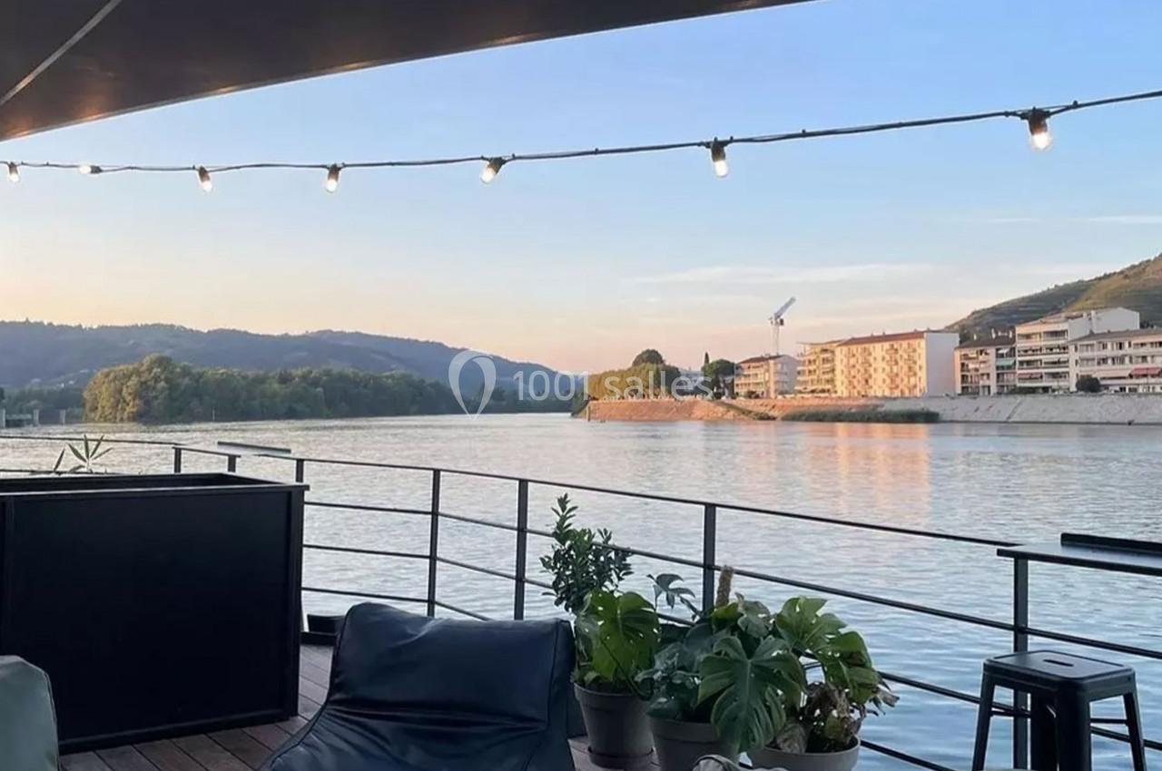 Vue depuis une terrasse en bois avec des plantes, donnant sur une rivière bordée de bâtiments et de collines au coucher du…