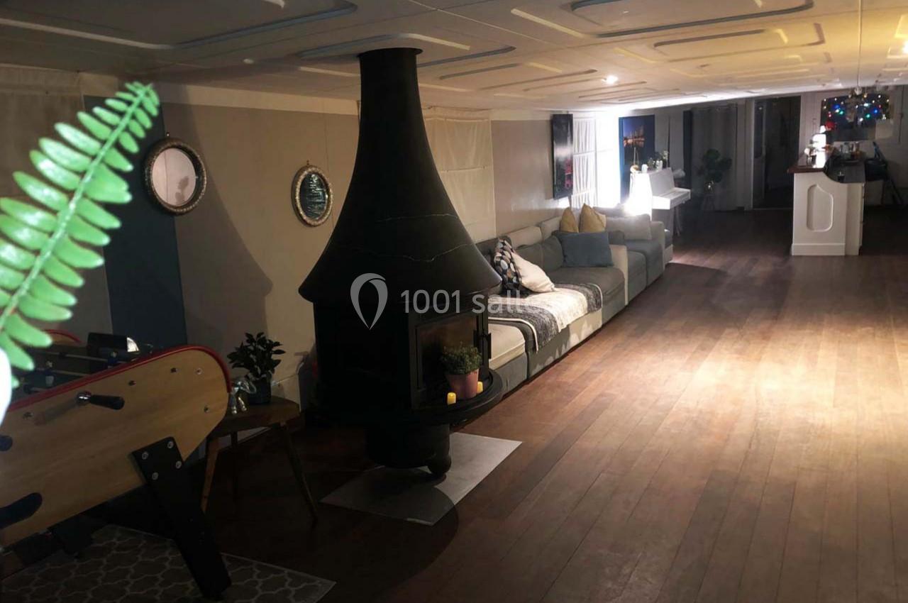 Salon spacieux avec poêle central, canapé gris, baby-foot et éclairage tamisé sur parquet en bois.