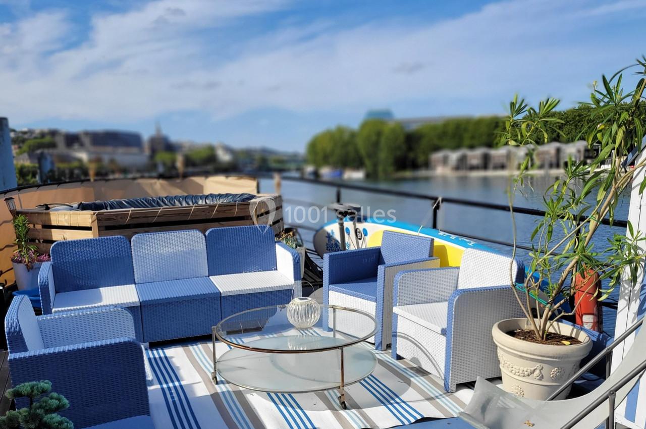 Salon de jardin avec canapés bleus et blancs sur une terrasse au bord de l'eau, entouré de plantes et d'une vue dégagée.