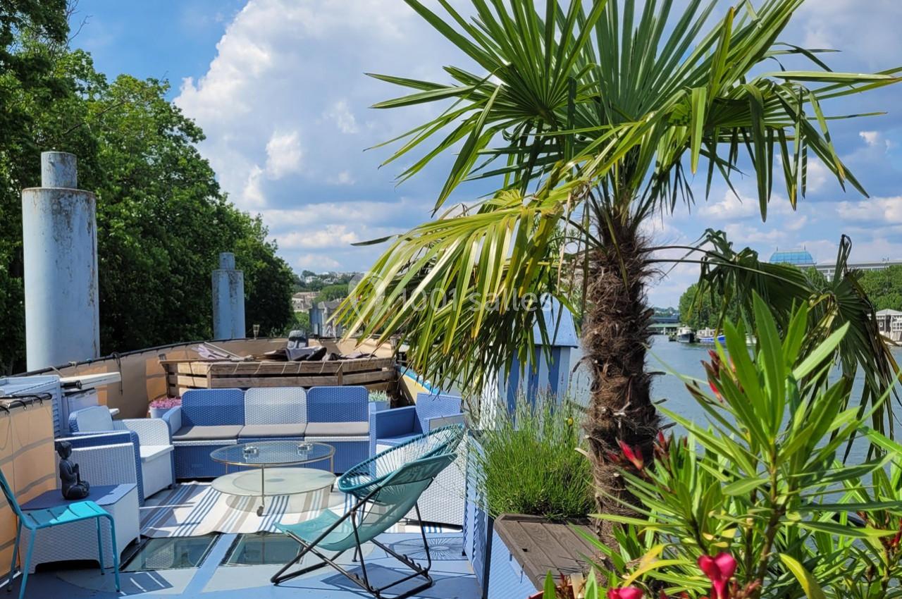 Terrasse aménagée sur une péniche avec fauteuils, table basse, plantes et vue sur un fleuve bordé de verdure.