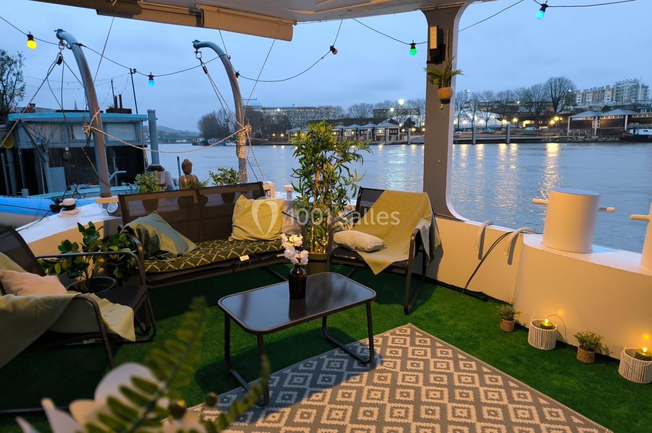 Terrasse aménagée sur une péniche avec mobilier de jardin, plantes et vue sur une rivière en arrière-plan.